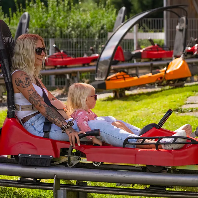 Eine Mutter mit Tattoos und Sonnenbrille sitzt mit ihrer Tochter in einem roten Rodelwagen auf der Grafenauer Sommerrodelbahn.