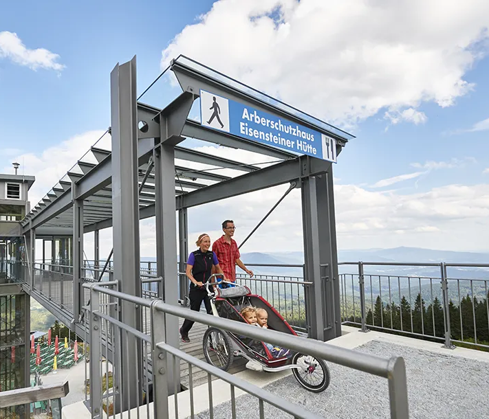 Familienausflug ohne Hindernisse mit atemberaubender Aussicht Auf der hohen modernen Aussichtsplattform mit Aufzug spaziert ein Paar mit einem Kinderwagen, während sich dünne Wolken über den Bergen formieren.
