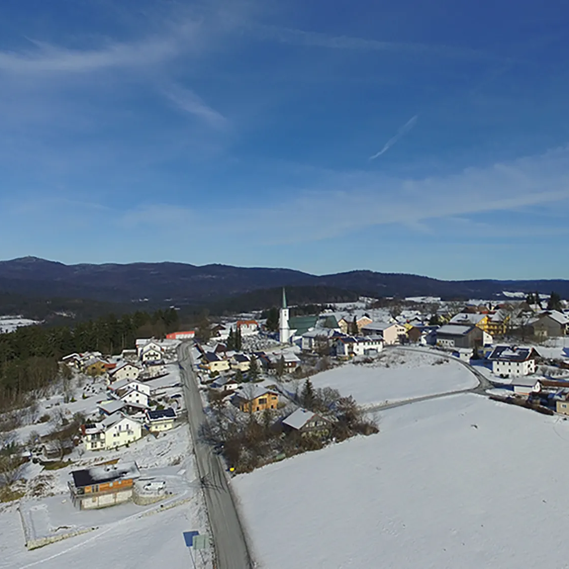 Das Dorf Hohenau in einer verschneiten Umgebung gelegen, umgeben von den Bayewaldbergen, mit der Kirche im Zentrum umrahmt von den anliegenden Häusern.