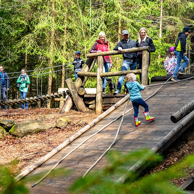 Spaß im Freien: Kinder auf der Holzrampe unterwegs am Naturspielplatz Ein Kind mit türkisfarbenem Shirt und bunten Stiefeln erklimmt die Holzrampe, umgeben von den Bäumen im Waldspielgelände unter der Beobachtung einer Gruppe von Erwachsenen.