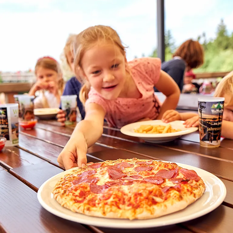 Ein Mädchen sitzt am Tisch und zieht einen Teller mit einer Salamipizza zu sich her. Kind greift nach Pizza