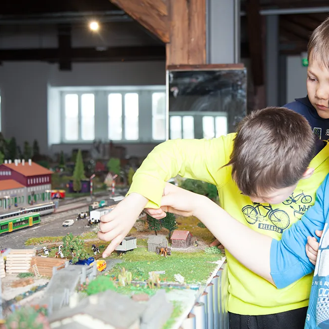 Kinder erkunden eine faszinierende Modellbahnlandschaft in den Naturparkwelten Bayerisch Eisenstein Die Jungen sind fasziniert von der Modelleisenbahn im Naturparkmuseum in Bayerisch Eisenstein, während sie auf die kleinen Figuren und Züge in der Anlage zeigen.