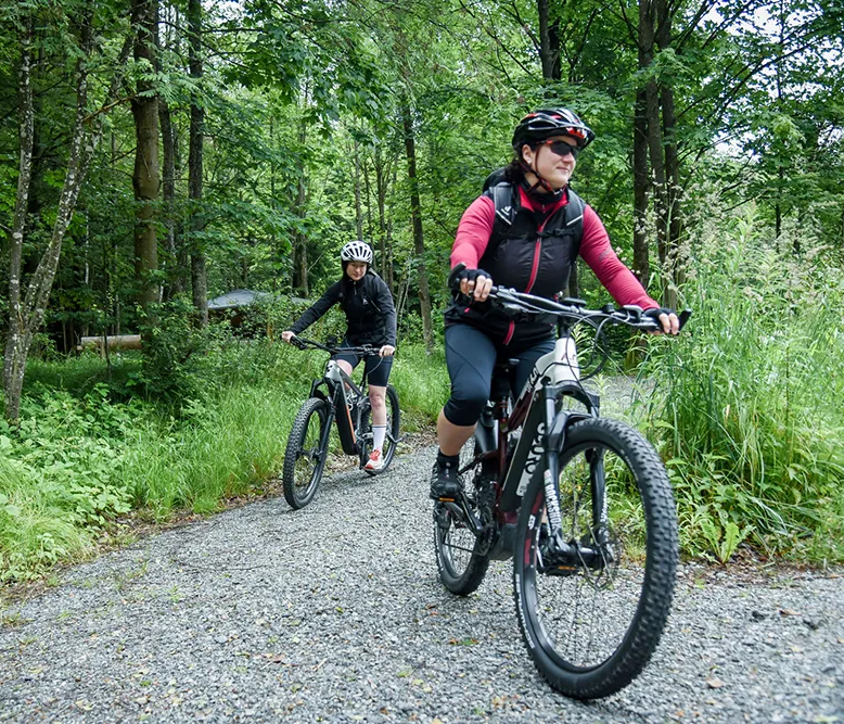 Zwei Radfahrerinnen fahren auf einem schmalen, kiesigen Waldweg