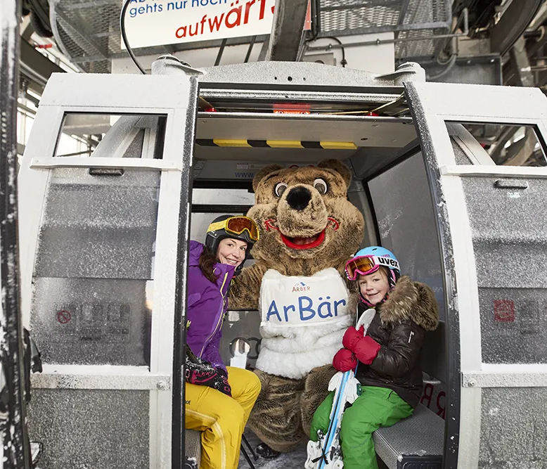 Spaßige Fahrt in der Arbergondel mit zwei Schwestern und dem Maskottchen ArBär In einer Gondel sitzen zwei Skifahrer-Mädels und ein Bärenmaskottchen, bereit um auf den Berg zu starten für ein Abenteuer im Schnee.