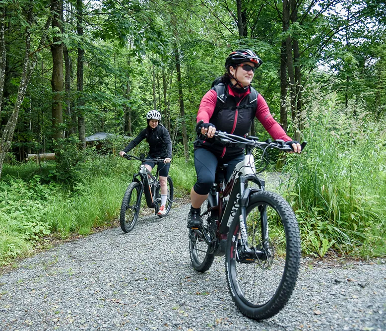 Zwei Radfahrerinnen fahren auf einem schmalen, kiesigen Waldweg