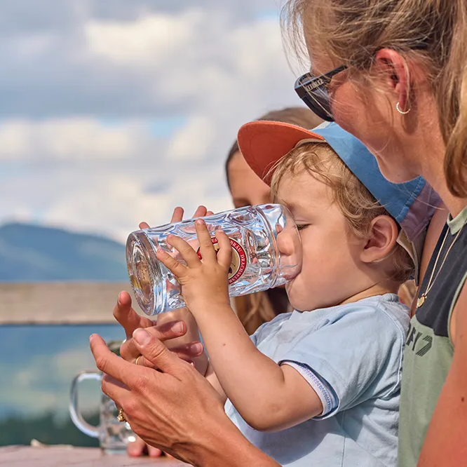 Glücksmomente: Kind erfrischt sich mit Getränk an einem Tisch im Freien Ein kleiner Junge stillt seinem Durst und trinkt aus einem großen Glas, während er von einer Frau mit langen Haaren umgeben ist, die ihn liebevoll unterstützt.