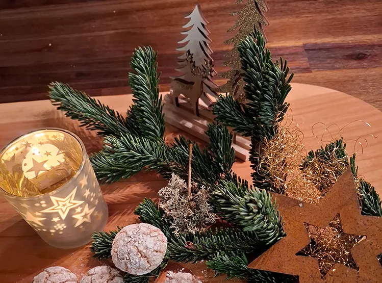 Ein rustikales Holzbrett mit festlichen kleinen Schokoladenbroten, Tannenzweigen und einer leuchtenden Kerze in einem Glas mit Sternen.