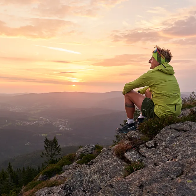 Entspannter Moment in der Natur bei Sonnenuntergang Die goldenen Sonnenstrahlen tauchen die Berge in warmes Licht, während ein Mann entspannt auf einem Felsen sitzt mit Blick in die Ferne.