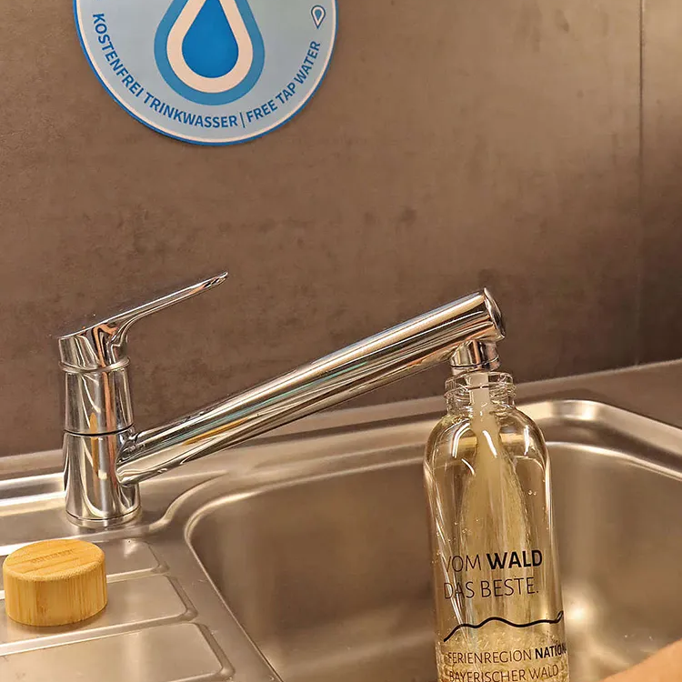 Der Wasserhahn sprudelt frisches Waldwasser in die nachhaltige Glasflasche, während ein Schild auf die kostenlose Trinkstation hinweist.