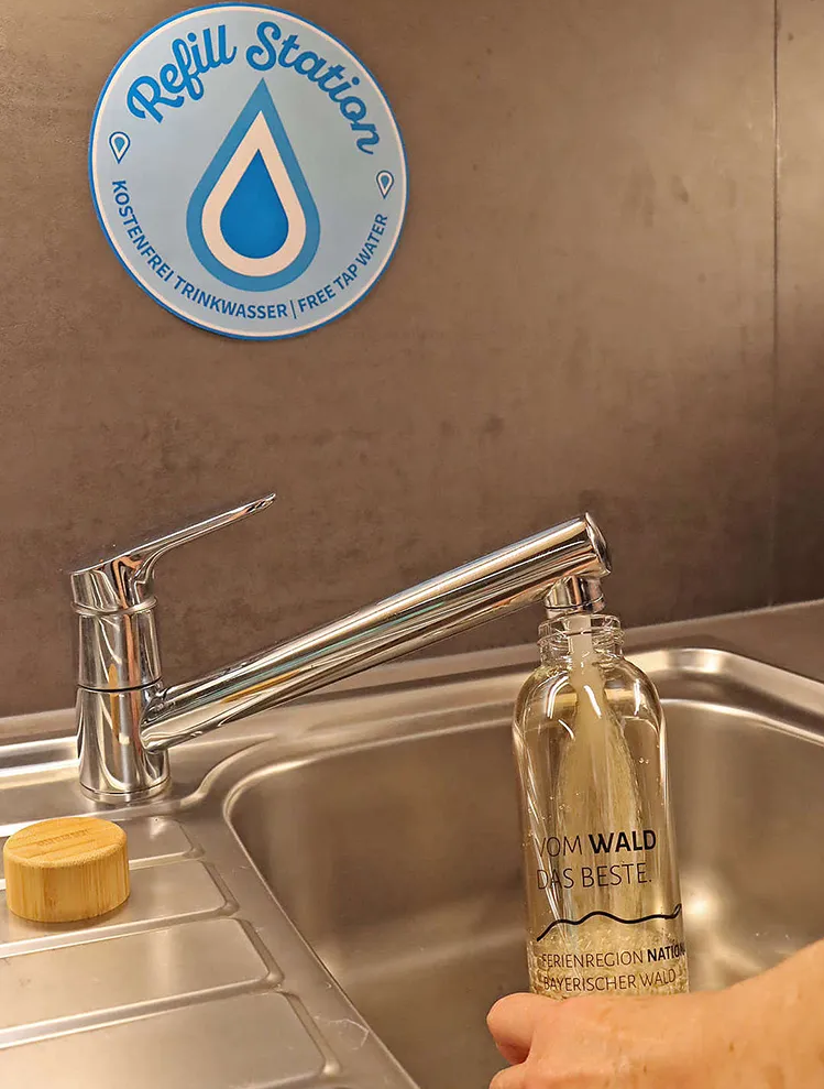 Wasserauffüllstation "Refill" für umweltfreundliche Trinkgewohnheiten Der Wasserhahn sprudelt frisches Waldwasser in die nachhaltige Glasflasche, während ein Schild auf die kostenlose Trinkstation hinweist.