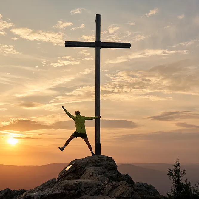 Lebensfreude und Freiheit auf dem Berggipfel erleben Vor einem imposante Sonnenuntergang steht eine Mann in Outdoorkleidung am Gipfelkreuz auf einem Felsen und hebt beschwingt einen Arm und ein Bein zur Seite.