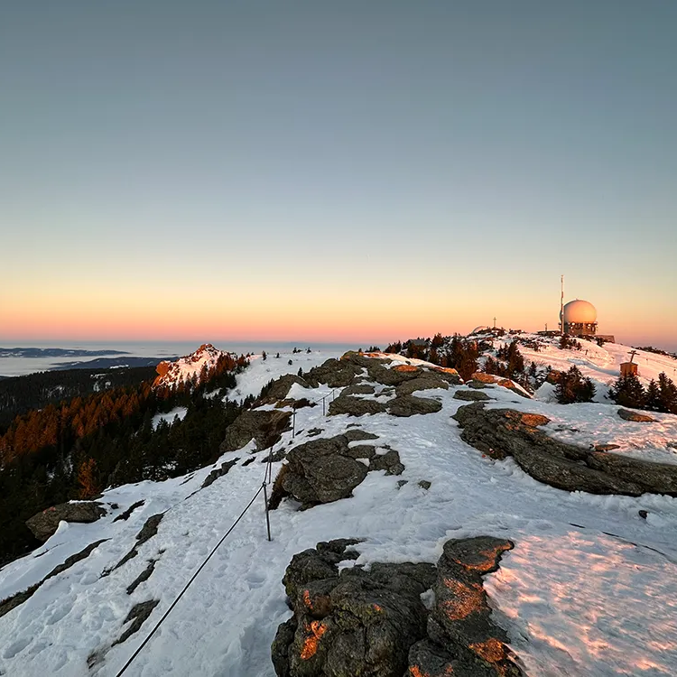 Winterlicher Anblick des Großen Arber Gipfels in der Dämmerung Der Sonnenuntergang färbt den Himmel über dem schneebedeckten Gipfel, während der Radarturm im Hintergrund sichtbar ist.