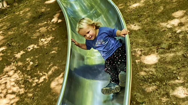 Ein kleiner Entdecker auf Rutschpartie am Waldspielgelände Ein kleines blondes Kind mit einem breiten Lächeln rutscht auf einer metallischen Rutsche, die auf einem Erdhügel angebracht ist.