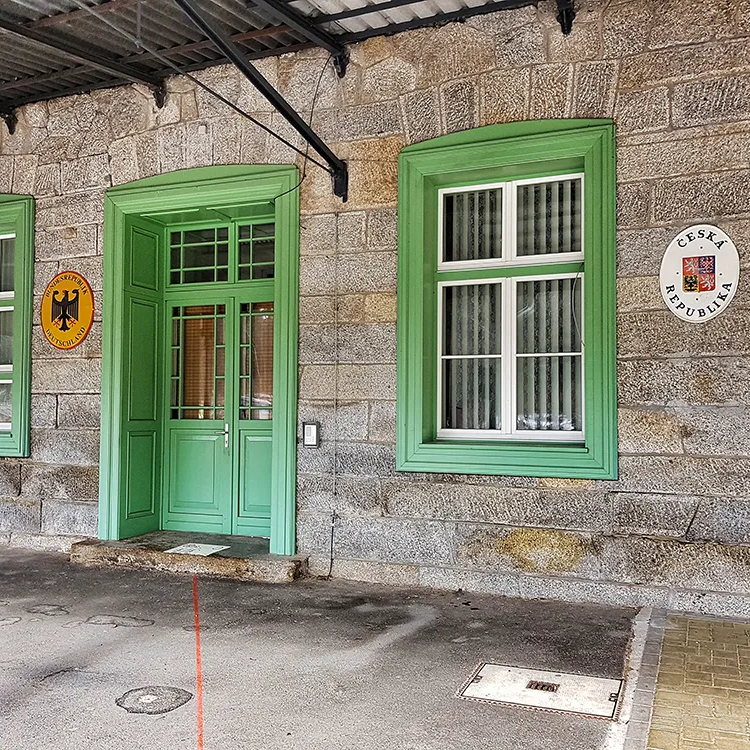 Historisches Bahnhofsgebäude am Grenzbahnhof in Bayerisch Eisenstein Das steinerne Gebäude des Bahnhofs Bayerisch Eisenstein mit grünen Fensterrahmen und einer markanten Eingangstür, geschmückt mit den Wappen Deutschlands und der Tschechischen Republik an der Wand..