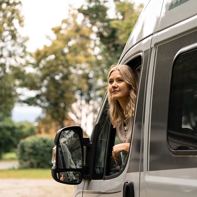 Entspanntes Reisen: Ein Blick aus dem Camper Eine blonde Frau genießt die Aussicht aus dem Fenster eines grauen Wohnmobils, während sie in der Natur parkt.