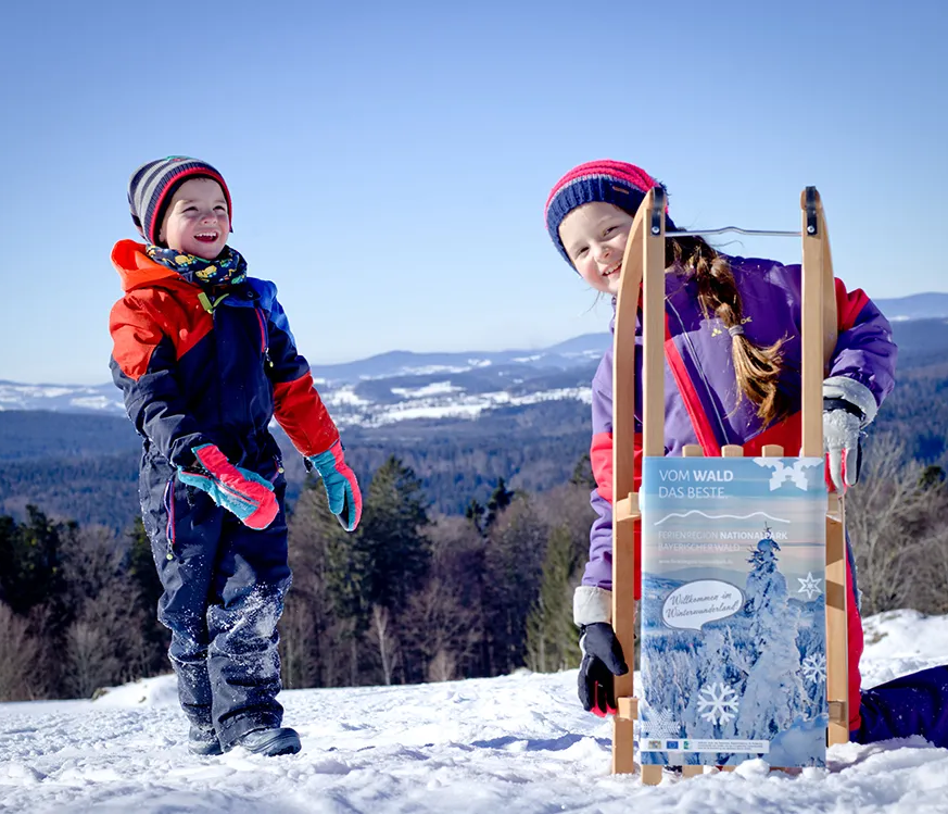 Kinder mit Rodelschlitten im Skigebiet Waldhäuser Zwei Kinder stehen fröhlich im Schnee, eines hält einen Schlitten mit einem Werbeschild für die Ferienregion Nationalpark Bayerischer Wald in den Händen