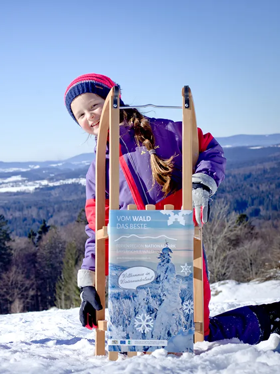 Kinder mit Rodelschlitten im Skigebiet Waldhäuser Zwei Kinder stehen fröhlich im Schnee, eines hält einen Schlitten mit einem Werbeschild für die Ferienregion Nationalpark Bayerischer Wald in den Händen