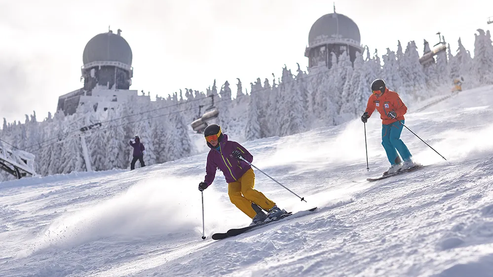 Skifahrer genießen den frischen Schnee im Arber-Skigebiet. Im Hintergrund die Arberkuppeln Zwei Skifahrer gleiten mit Schwung die verschneite Piste am Großen Arber hinunter. Im Hintergrund die Radarkuppeln.