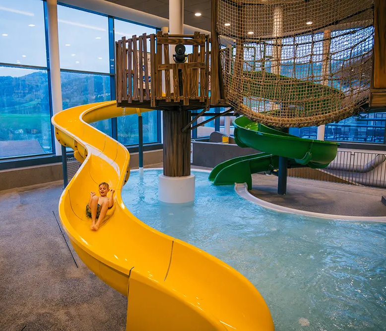 Wasservergnügen: Kind rutscht fröhlich ins Wasser Im Hallenbad des Schreinerhofes, rutscht ein Kind mit Begeisterung die gelbe Rutsche hinunter, während die Landschaft draußen sichtbar ist.