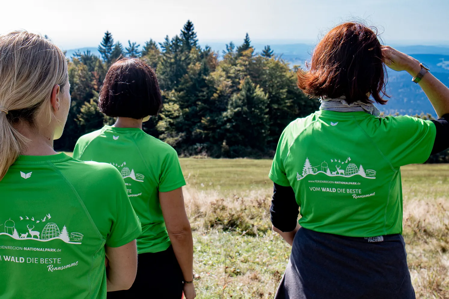 Die Mitarbeiterinnen der Ferienregion tragen neongrüne T-Shirt im "Vom Wald das Beste"-Design mit dem FNBW-Logo und genießen die Aussicht auf die hügelige, bewaldete Umgebung.