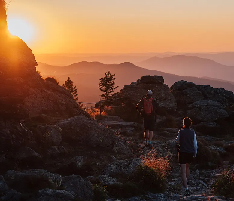 Romantische Abendwanderung mit atemberaubendem Ausblick vom Arber Der Mann mit Rucksack und die Frau, nur noch in schattenhaften Silhouetten, wandern den felsigen Weg hinauf, während die Sonne eine goldene Abendstunde schafft.