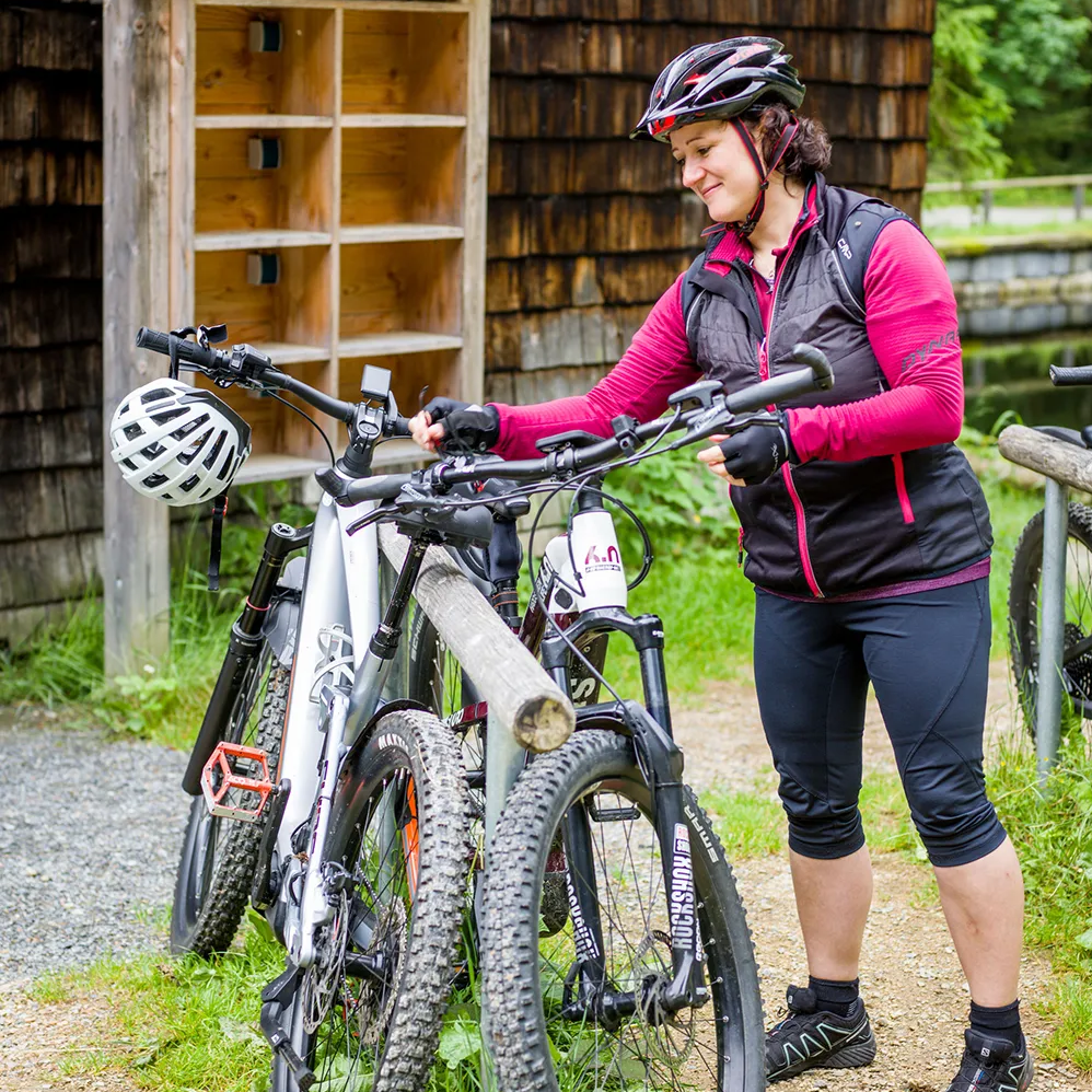 Fahrradparkplatz in der malerischen Waldlandschaft am Rachelradweg Eine Radfahrerin in sportlicher Kleidung überprüft ihr Mountainbike, das Sie an einem Holzständer vor der Racheldiensthütte abgestellt hat.