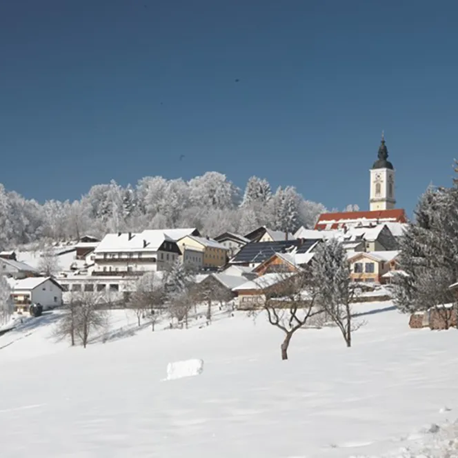 Kirchdorf im Wald im Winter, mit schneebedeckten Dächern und einer markanten Kirchturmspitze, die in den blauen Himmel ragt.