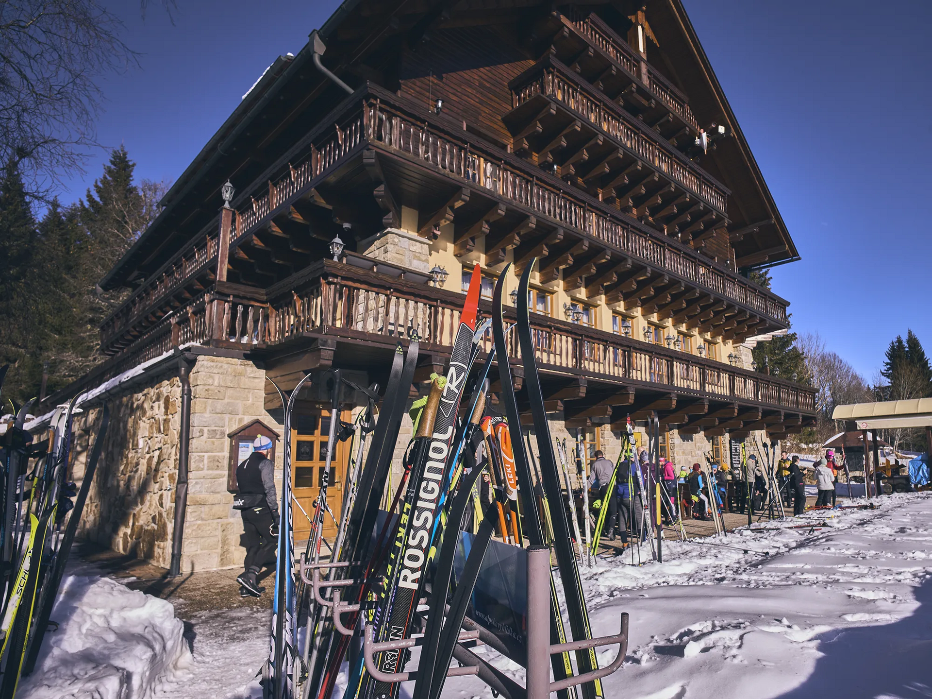 Skifahrer genießen den Tag vor einer Berghütte Das Bild zeigt ein rustikales Skihütte mit Holzverkleidung, umgeben von Schnee und Langläufern, die einen Langlauftag an der Grenze zum Sumava genießen.