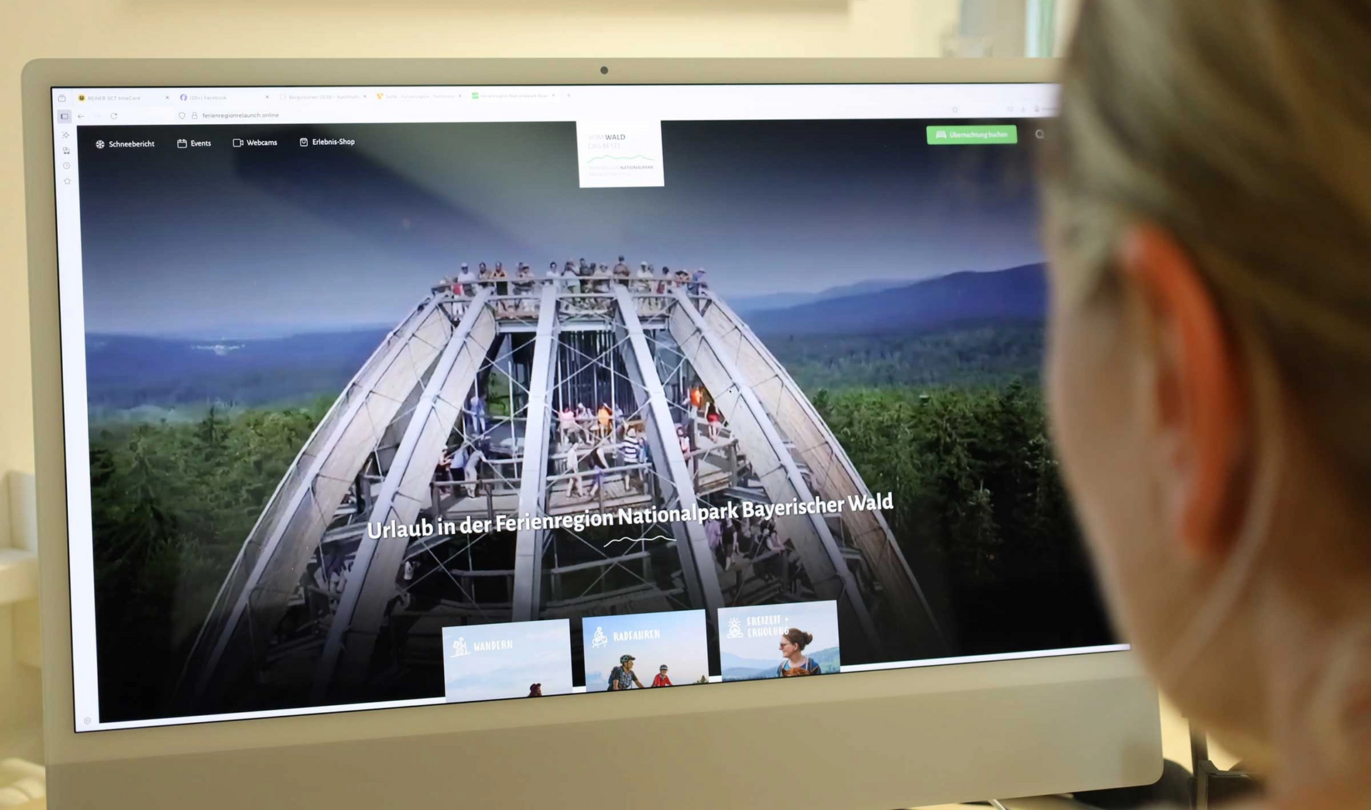 Frau plant ihren Urlaub am Laptop in einem hellen Raum Eine Frau mit blondem Zopf sitzt vor einer Bildschirm Anzeige mit dem Baumwipfelpfad Bayerischer Wald in Neuschönau, einer Gemeinde der Ferienregion.