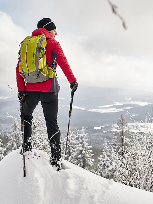 Winterwanderer genießt die Aussicht auf die Winterlandschaft in der Ferienregion Bayerischer Wald Ein Wanderer in roter Jacke und grünem Rucksack steht auf einem schneebedeckten Felsen und blickt auf die winterliche Landschaft der Ferienregion Nationalpark Bayerischer Wald