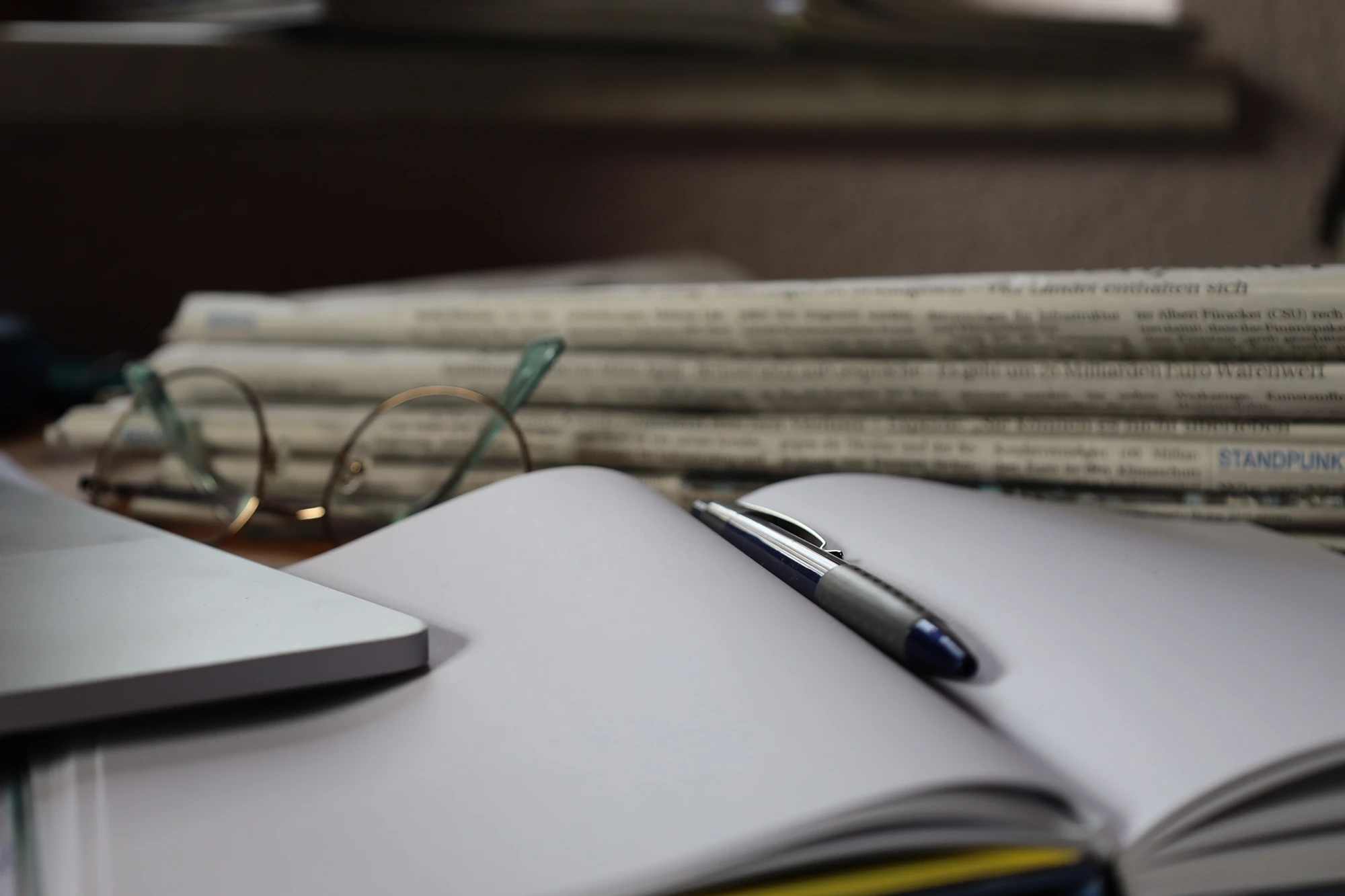 Ein Stapel Zeitungen liegt auf einem Tisch, daneben ein aufgeschlagenes Notizbuch mit Stift und eine Brille.